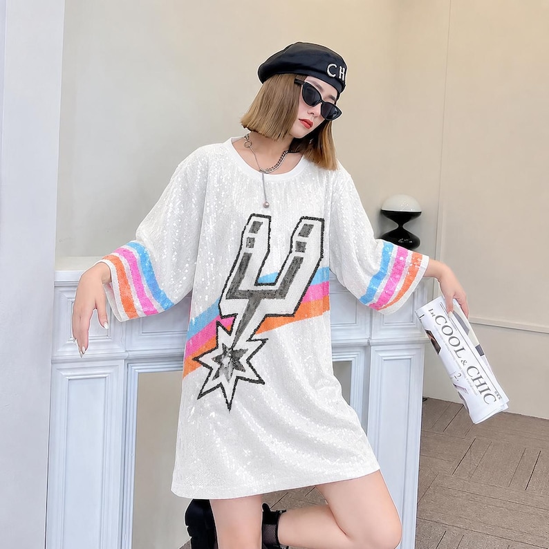 May include: White sequined dress with a graphic design featuring a star and lightning bolt. The sleeves have rainbow stripes. The dress is knee-length and has a loose fit. The model is wearing a black beret and sunglasses.