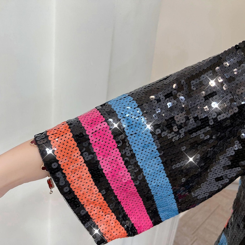 May include: Close-up of a black sequined sleeve with colorful stripes. The stripes are orange, pink, and blue. The sequins sparkle, reflecting light. The sleeve is part of a garment.
