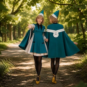 May include: Two people wearing teal dresses with matching capes and hats. The dresses have black accents and white underskirts. The capes feature a white design on the back. Both are wearing yellow heels and black tights. The background is a path through a wooded area.