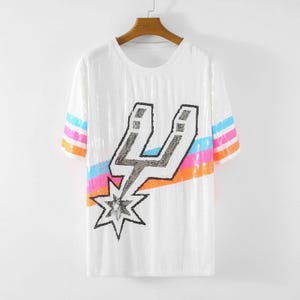 May include: White sequined t-shirt with a crew neck and short sleeves. The shirt features a large black and silver graphic of a stylized letter "U" with a starburst design. The sleeves have horizontal stripes in blue, pink, and orange.