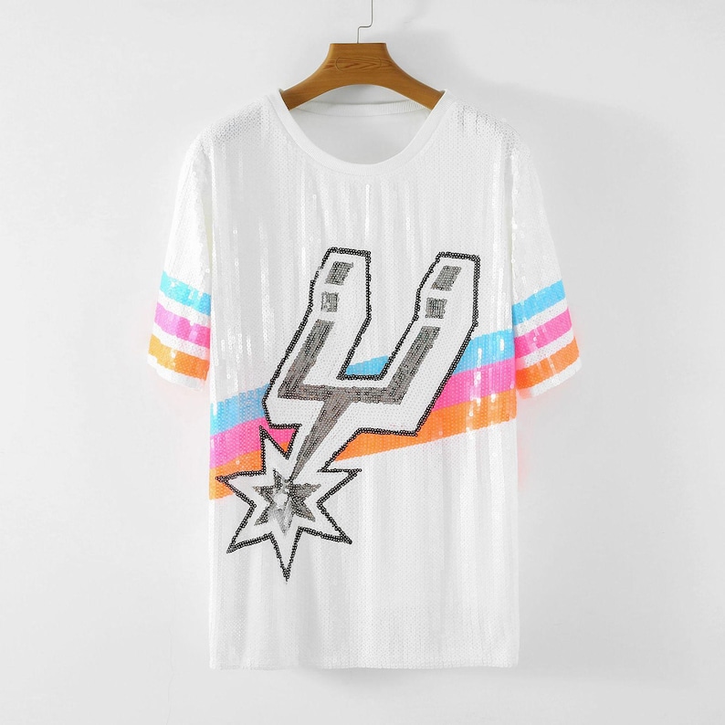 May include: White sequined t-shirt with a crew neck and short sleeves. The shirt features a large black and silver graphic of a stylized letter "U" with a starburst design. The sleeves have horizontal stripes in blue, pink, and orange.