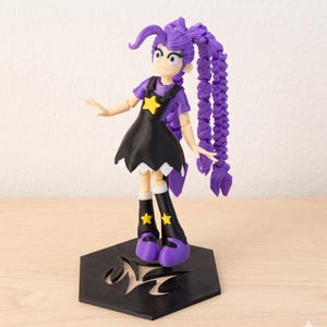 以下が含まれることがあります： 紫色の髪、黒いドレス、星のアクセントが特徴のアニメ風フィギュア。フィギュアは、様式化されたデザインの黒い六角形のベースに立っています。紫色のトップス、黄色の星が付いた黒いブーツ、紫色の靴を履いています。
