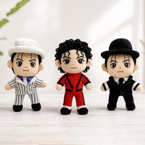 Puede incluir: Tres muñecos de peluche de una estrella del pop, cada uno con un atuendo diferente. Uno lleva un traje y sombrero de rayas blancas, otro un mono rojo y el tercero un traje y sombrero negros. Todos los muñecos tienen los mismos rasgos faciales.