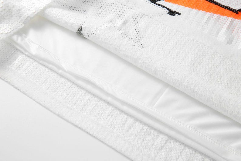 May include: Close-up of a white sequined fabric with an orange and black design. The fabric is layered over a white lining. The sequins create a textured surface, reflecting light.