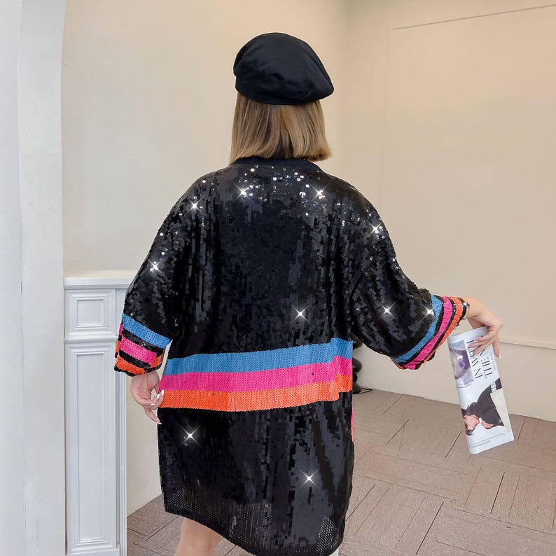 May include: Black sequin kimono-style jacket with horizontal stripes of pink, blue, and orange. The sleeves have matching stripes. A black beret is worn on the head. The jacket is knee-length.