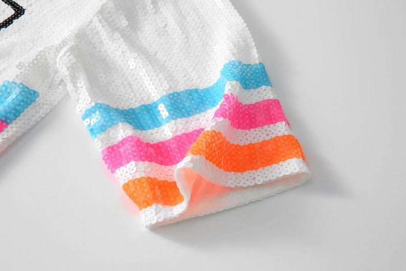 May include: White sequined fabric with colorful horizontal stripes in blue, pink, and orange. The fabric appears to be part of a garment, possibly a sleeve or trim, with a bright and playful design.