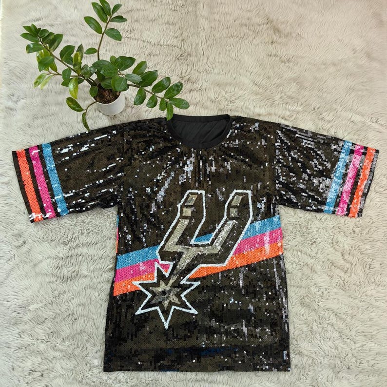 May include: Black sequined t-shirt with a colorful design. The shirt features a silver and white lightning bolt and star design, with a blue, orange, and pink stripe. The sleeves have matching stripes.