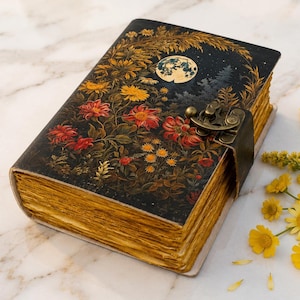 May include: A rectangular journal with a dark cover featuring a full moon, pine trees, and vibrant red and yellow flowers. The book is secured with a metallic latch and has thick, deckled-edge pages with a golden-yellow hue throughout the paper.