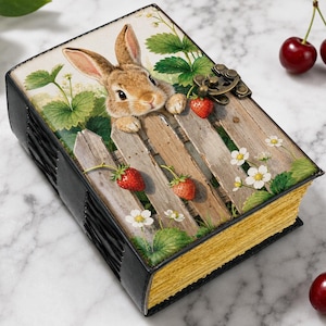 May include: A rectangular journal with a black spine and gold-toned page edges. The cover features a brown rabbit peeking over a wooden fence surrounded by green leaves, white flowers, and red strawberries. A metal latch secures the book closed.
