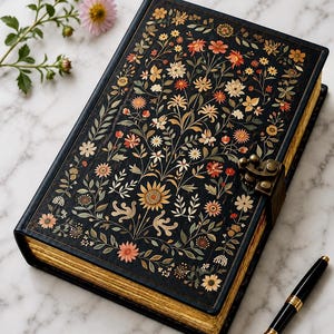 Puede incluir: Un diario de tapa dura negra con un patrón floral detallado en tonos naranja, amarillo y verde. El libro tiene bordes de página dorados y un cierre de pestillo metálico. Una pluma estilográfica negra y dorada se sitúa junto al diario.