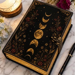 May include: A black hardcover journal featuring a gold-toned moon phase illustration on the front cover, surrounded by intricate floral patterns. The book includes a metallic latch closure and gold-gilded page edges, accompanied by a black ballpoint pen.