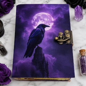 May include: A rectangular journal featuring a purple cover with a graphic print of a black raven perched on a spire against a full moon and cloudy sky. The journal includes a decorative antique brass-colored metal latch closure on the right side.