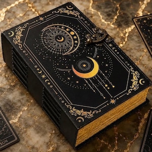 May include: A black rectangular journal with gold celestial motifs including moons and stars on the cover, featuring a metal latch closure and gold-edged pages, accompanied by three tarot cards labelled The Sun, The Moon, and XVII.