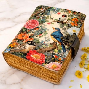 Könnte beinhalten: Ein dekoratives Notizbuch mit Blumen- und Vogeldesign. Das Cover zeigt eine lebendige Illustration von Vögeln, die auf Ästen inmitten bunter Blumen sitzen. Das Notizbuch hat einen Vintage-Look mit einem Metallverschluss und gealterten Seiten.