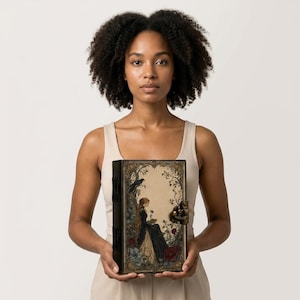 K&ouml;nnte beinhalten: Ein rechteckiges Hardcover-Buch mit einer detaillierten Illustration einer Person in einem langen dunklen Kleid, die unter einem Baum mit einem Raben sitzt. Der Einband hat verzierte florale R&auml;nder, einen Metallverschluss und einen dunklen Buchr&uuml;cken.