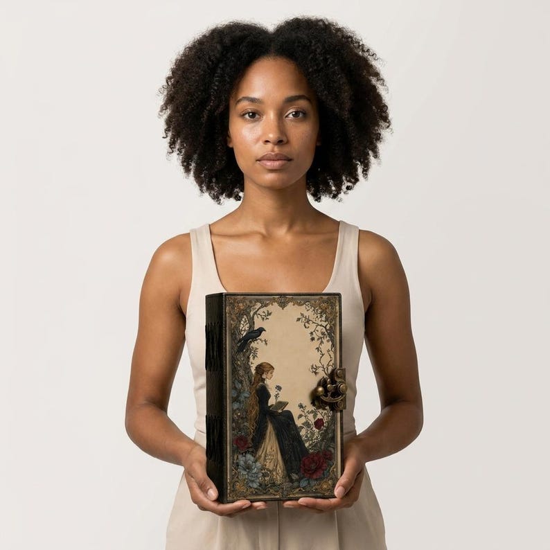 K&ouml;nnte beinhalten: Ein rechteckiges Hardcover-Buch mit einer detaillierten Illustration einer Person in einem langen dunklen Kleid, die unter einem Baum mit einem Raben sitzt. Der Einband hat verzierte florale R&auml;nder, einen Metallverschluss und einen dunklen Buchr&uuml;cken.