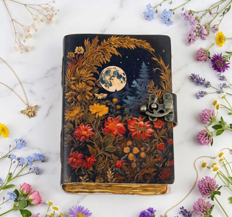 May include: A rectangular journal with a dark blue cover featuring a full moon, pine trees, and various red, orange, and yellow flowers. The journal has a metallic antique-style latch closure and deckled, aged-looking paper edges.