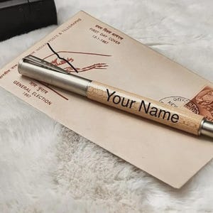 May include: A ballpoint pen with a light-colored wooden barrel and silver-toned metal accents, including a clip and end caps. The words "Your Name" are printed in a dark, sans-serif font on the wooden section of the pen body.