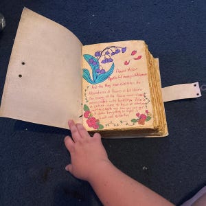 K&ouml;nnte beinhalten: Ein handgefertigtes Tagebuch mit einem hellbraunen Lederumband und Riemenverschluss. Die offene Seite zeigt eine handgezeichnete Illustration von violetten Maigl&ouml;ckchen, roten Rosen und orangefarbenen Bl&uuml;ten sowie handschriftlichen Text zum Flower Moon.