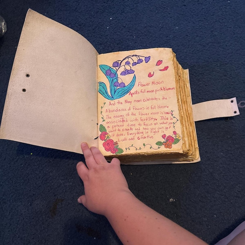 K&ouml;nnte beinhalten: Ein handgefertigtes Tagebuch mit einem hellbraunen Lederumband und Riemenverschluss. Die offene Seite zeigt eine handgezeichnete Illustration von violetten Maigl&ouml;ckchen, roten Rosen und orangefarbenen Bl&uuml;ten sowie handschriftlichen Text zum Flower Moon.