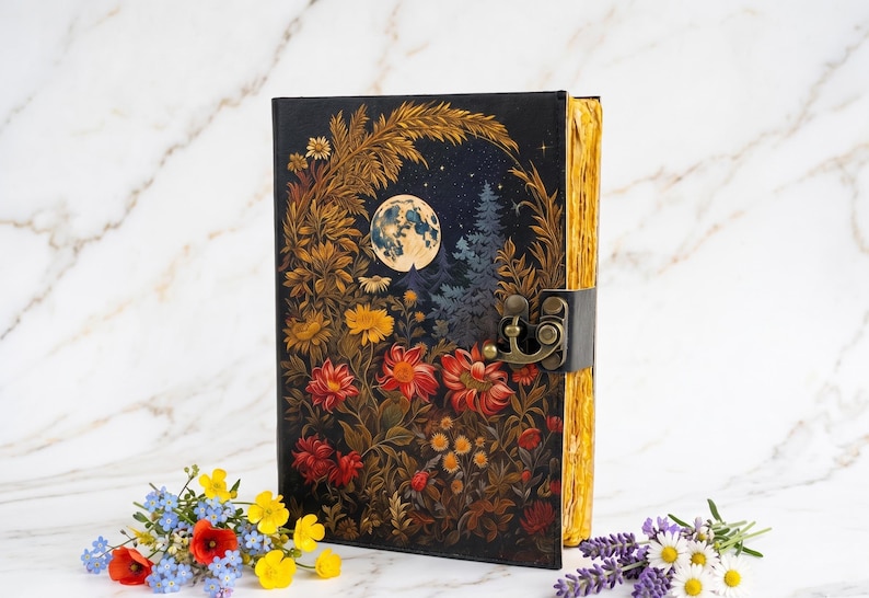 May include: A black rectangular journal featuring a full-color illustration of a night scene with a full moon, pine trees, and vibrant red, orange, and yellow flowers. The book has a metallic bronze-colored latch and gold-toned page edges.
