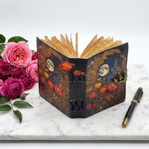 May include: A journal with a dark cover featuring a floral and moon print, bound with black stitching and a metal latch. The pages are deckled and light brown. A black fountain pen lies nearby, and a cluster of pink roses is positioned to the left.