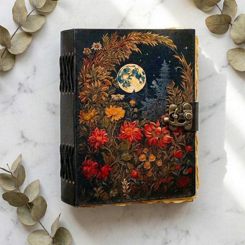 May include: A rectangular journal with a black cover featuring a vibrant illustration of a full moon, pine trees, and colorful red, orange, and yellow flowers. The book has a dark metal latch closure on the right side and visible black binding stitches.