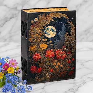 May include: A black hardcover journal featuring a full-color illustration of a night scene with a large moon, pine trees, and vibrant red, yellow, and orange flowers. The book has a metal latch closure and black cord lacing along the spine.