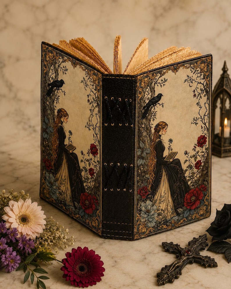 K&ouml;nnte beinhalten: Ein Notizbuch mit festem Einband und schwarzem Buchr&uuml;cken mit Kreuzstich-Details. Vorder- und R&uuml;ckseite zeigen eine Illustration einer Person mit langem Haar in einem dunklen Kleid, die neben einem Baum mit Raben und Blumen sitzt.