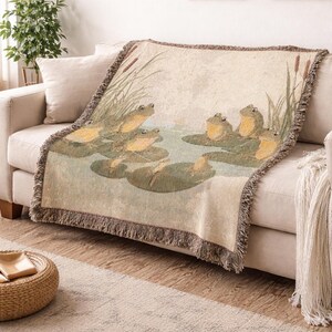 May include: A woven throw blanket depicting a pond scene with frogs on lily pads. The blanket has a beige background with a detailed illustration in shades of green, yellow, and brown. The edges are finished with a fringed border.