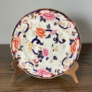 May include: A round ceramic plate featuring a cream-colored base with a vibrant floral pattern in shades of pink, orange, and yellow. The design includes deep blue scrolling accents and a scalloped edge trimmed with a thin gold-colored border.