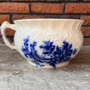 May include: Cream-colored antique chamber pot with blue floral designs. The pot has a handle and a gold trim around the base. The background is a brick wall.