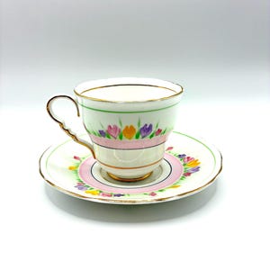 May include: A white porcelain teacup and saucer set featuring a delicate floral pattern with pink, yellow, and purple flowers. The rim of the cup and saucer are accented with gold trim, and the handle has a scalloped, wavy edge design.
