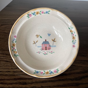 May include: Cream-colored ceramic bowl with a floral border and a central illustration of a pink house with a blue roof, a white picket fence, and butterflies. The bowl has a scalloped edge and is suitable for serving soups or salads.