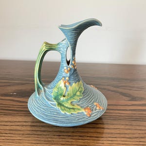 May include: A blue ceramic pitcher with a textured surface, featuring a green leaf and orange berry relief design. The pitcher has a curved handle and a flared spout, with a distinct opening between the handle and the neck.