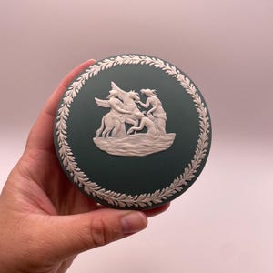 May include: A round, dark green jasperware trinket box lid featuring a white relief design of three figures and a winged horse. The lid is decorated with a white laurel leaf border encircling the central mythological scene in matte finish.