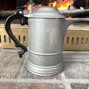 May include: A vintage silver-colored metal coffee pot with a black handle and lid. The pot has a spout and is sitting on a brick surface. The background shows a fireplace with burning wood.