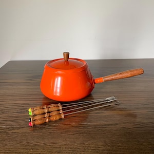 May include: A bright orange fondue pot with a matching lid, a wooden handle, and a wooden lid knob. The set includes four metal fondue forks with carved wooden handles, each featuring a small colored tip in yellow, green, black, or red.