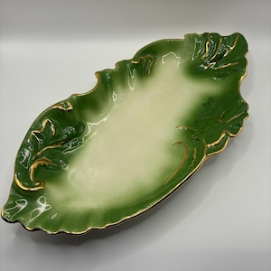 May include: An oval ceramic serving dish with a cream-colored center and a deep green border. The edges are shaped like stylized leaves with scalloped rims, and the entire perimeter is accented with a thin, metallic gold painted trim.