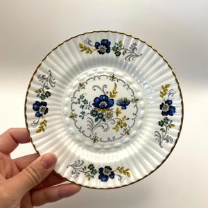 May include: White porcelain saucer with a fluted design and a gold rim. The saucer is decorated with blue and yellow floral patterns. A hand is holding the saucer.