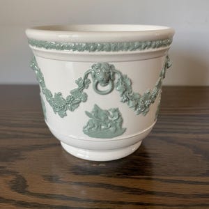 May include: White ceramic planter with a light green decorative design. The planter features a detailed relief pattern of a garland, cherubs, and lion heads. The rim and handles are also decorated with the same light green color.