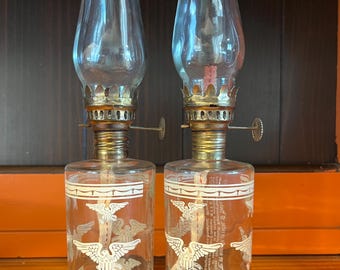 May include: Two clear glass oil lamps with a white eagle design. The lamps have a clear glass chimney, a brass-colored metal base, and a wick. The lamps are likely vintage or antique.