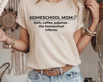 May include: A beige t-shirt with the text "HOMESCHOOL MOM: faith, coffee, pajamas - the homeschool trifecta." The shirt has short sleeves and is paired with denim shorts and a brown belt.