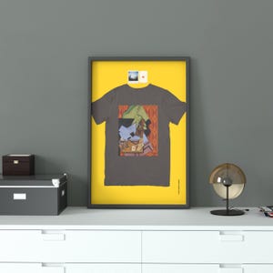 Può includere: Un poster incorniciato che mostra una t-shirt grigio scuro centrata su uno sfondo giallo brillante. La t-shirt presenta una stampa grafica in stile cubista con toni tenui di verde, blu e arancione. Due piccole foto quadrate sono appuntate sopra.