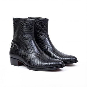 Può includere: Un paio di stivaletti in pelle nera con una texture a pelle di serpente. Gli stivaletti hanno la punta affusolata, un piccolo tacco e un dettaglio con fibbia laterale. Gli stivaletti sono alti circa 15 cm.