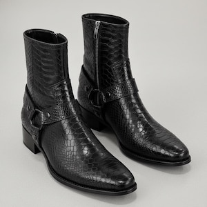 Könnte beinhalten: Ein Paar schwarze Leder-Ankle-Boots mit Schlangenleder-Textur. Die Stiefel haben eine spitze Zehenpartie, einen seitlichen Reißverschluss und einen Riemen mit Metallschnalle. Die Stiefel haben einen niedrigen Absatz.