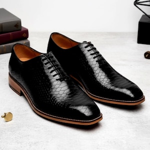 Puede incluir: Un par de zapatos de vestir de cuero negro con un patrón texturizado similar a la piel de reptil. Los zapatos tienen un diseño Oxford clásico con cordones negros, suela marrón claro y tacón de madera. Se ve un gemelo dorado.
