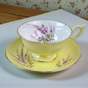 May include: A yellow teacup and saucer set with a floral design. The teacup and saucer are yellow with a gold trim. The inside of the teacup is white with a purple and green floral pattern. The saucer has a matching floral pattern.