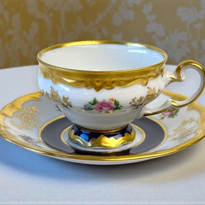以下が含まれることがあります： 金色の縁取りと花のアクセントが付いた白いティーカップとソーサーのセット。カップとソーサーは金色の縁とネイビーブルーと金のベースが付いています。セットはピンクと黄色のバラで飾られています。