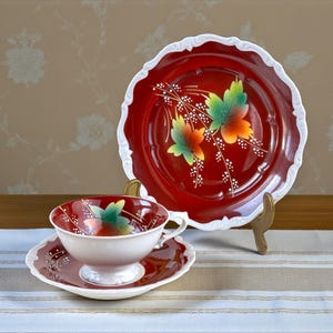 Pode incluir: Conjunto de chá decorativo vermelho e branco. O conjunto inclui uma chávena, um pires e um prato grande. As peças vermelhas têm uma borda branca ondulada e um desenho floral com folhas verdes, laranja e amarelas. Sobre uma toalha de mesa listrada.